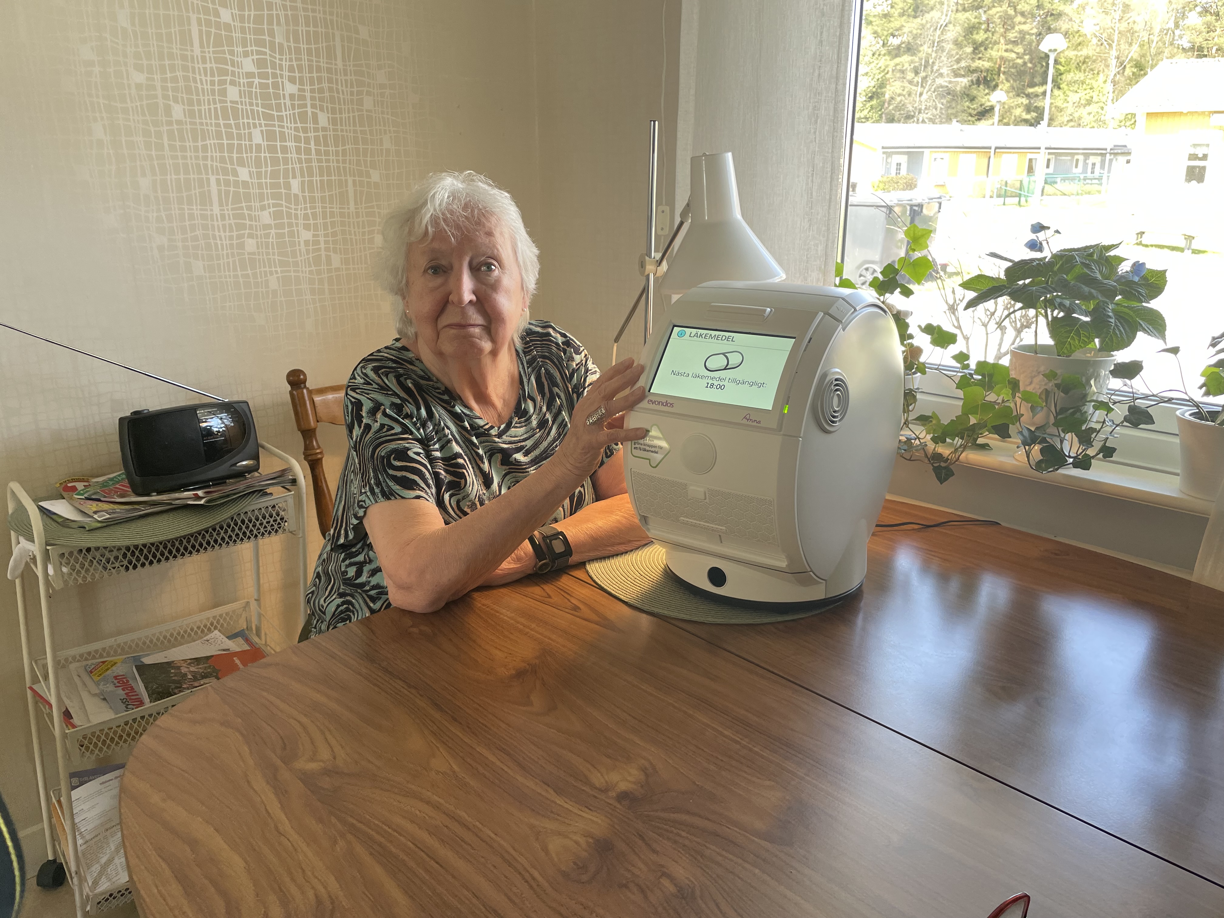 Äldre kvinna sitter vid sitt köksbord bredvid hennes medicingivare.