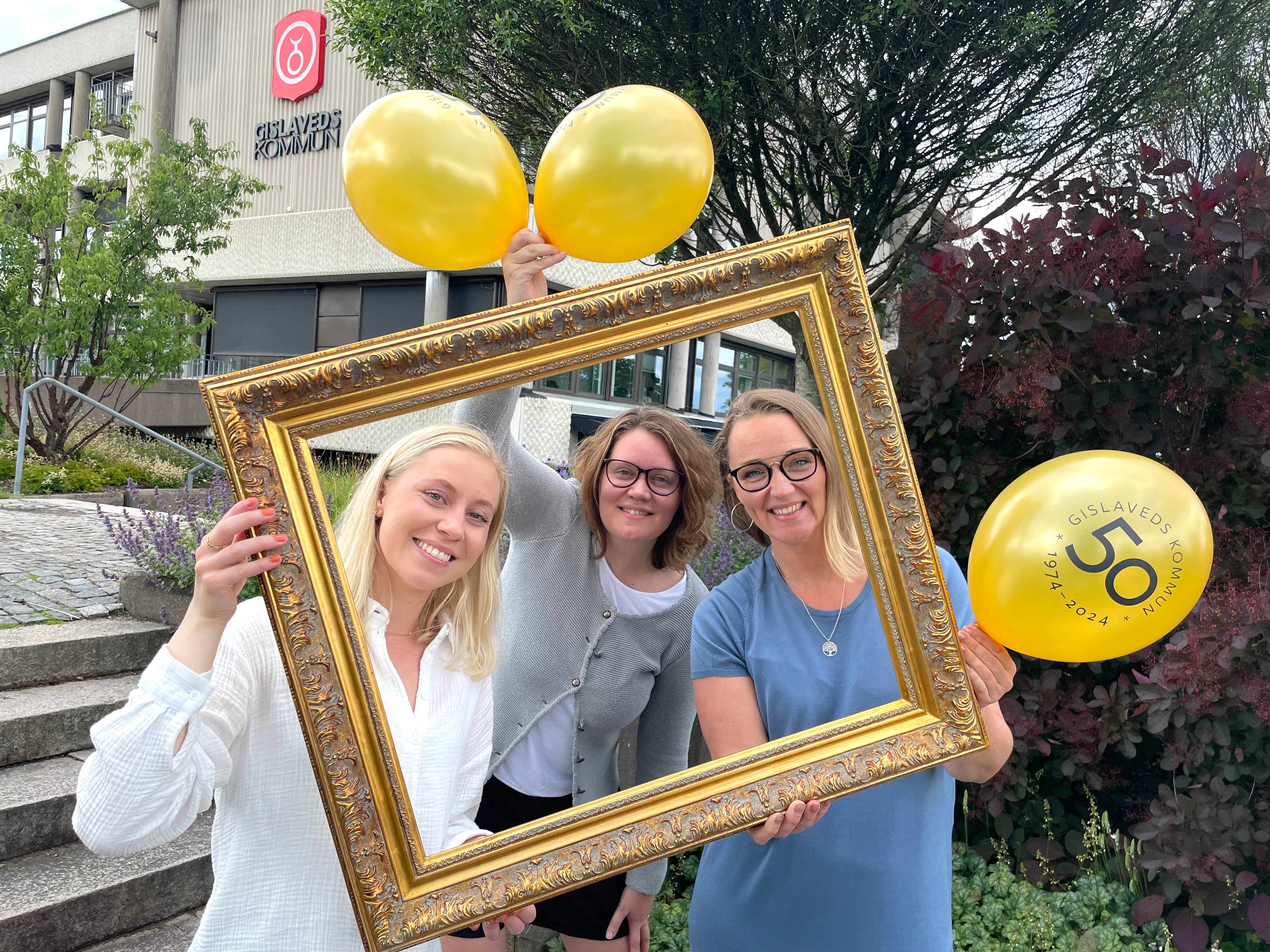 Tre kvinnor tittar fram bakom en guldig ram med ballonger i händerna.