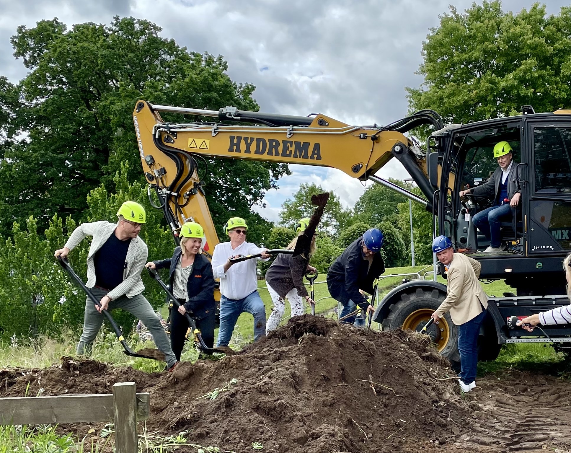 ett antal personer tar ett symboliskt första spadtag ur en jordhög. I bakgrunden sitter en man i en grävskopa.