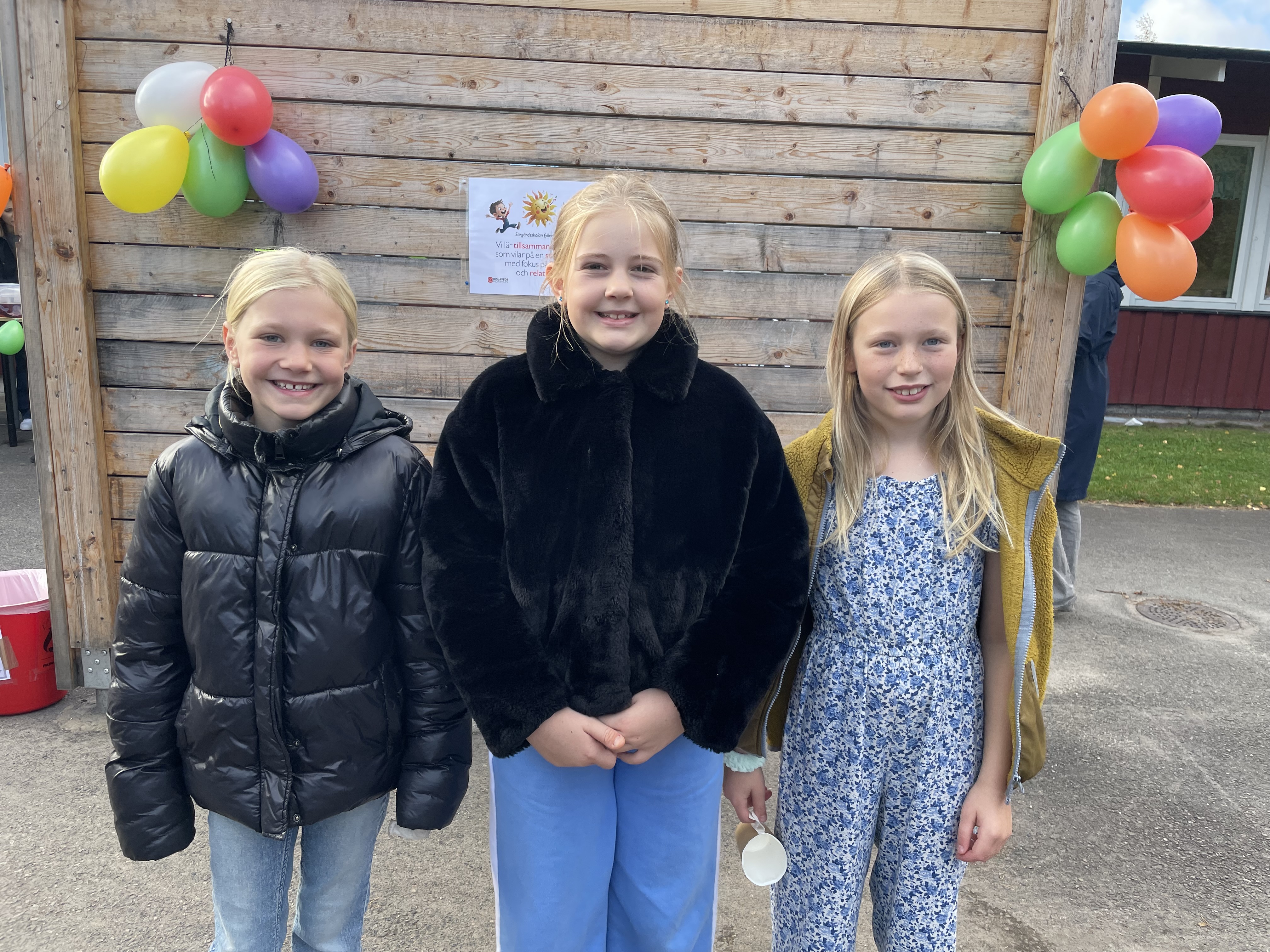 Tre tjejer i 10-årsåldern står framför en vägg med ballonger på.