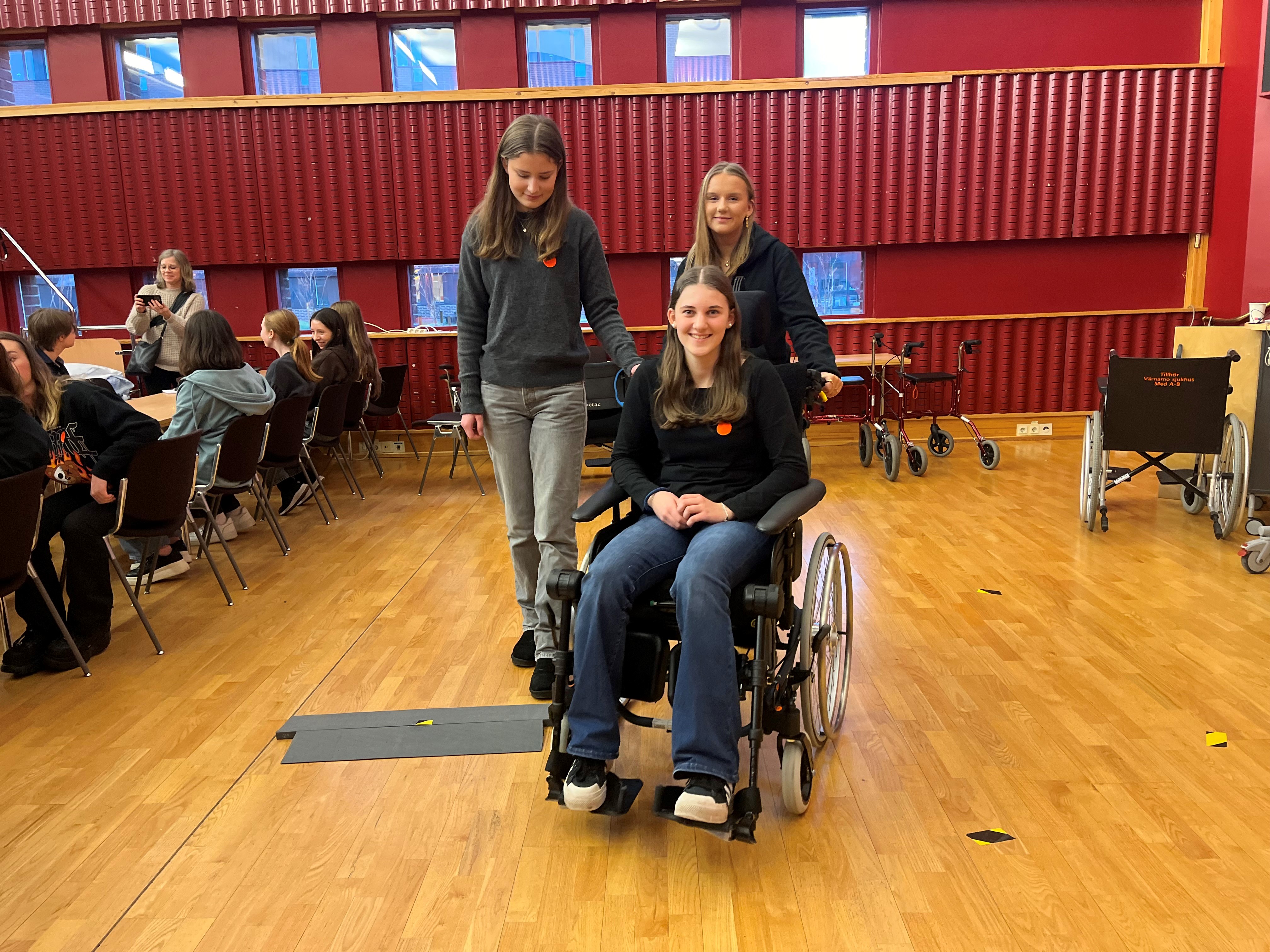 En tjej provar att sitta i en rullstol och köra över ett litet gupp. Bakom henne står två andra tjejer.