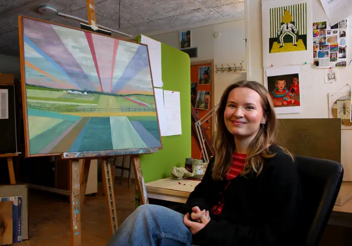 En kvinna sitter i en fåtölj framför ett staffli med en målning.