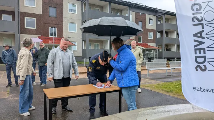Representanter från olika aktörer står i kö till ett bord där avtalet skrivs under. Bordet står utomhus där det både regnar och blåser.