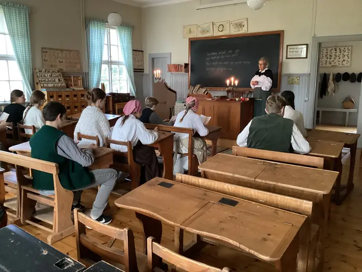 Ett foto som visar en gammaldags skolsal med elever och en fröken vid katedern.