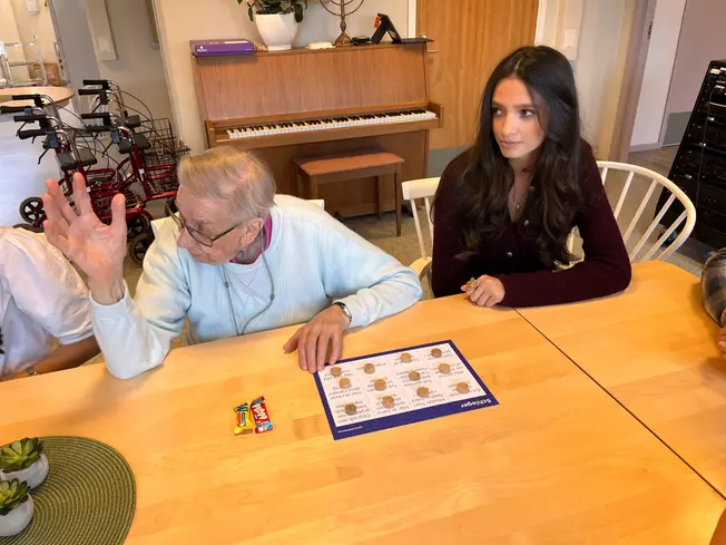 En äldre dam räcker upp handen. En ung tjej sitter bredvid.