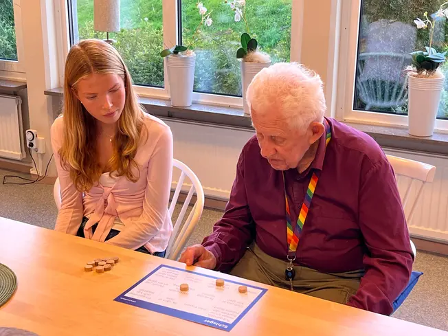 En äldre herre tittar på sin bingo-bricka tillsammans med en elev.