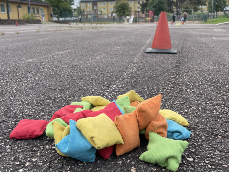 Ärtpåsar i olika färgen ligger på asfalten. I bakgrunden syns en röd kon.