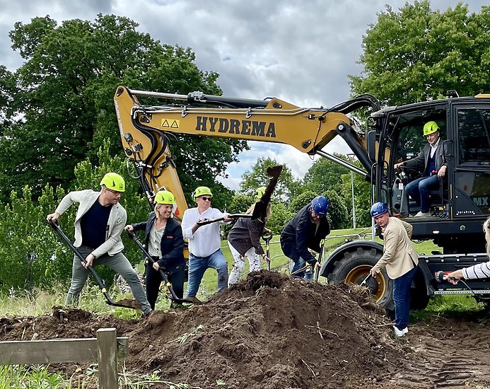 ett antal personer tar ett symboliskt första spadtag ur en jordhög. I bakgrunden sitter en man i en grävskopa.