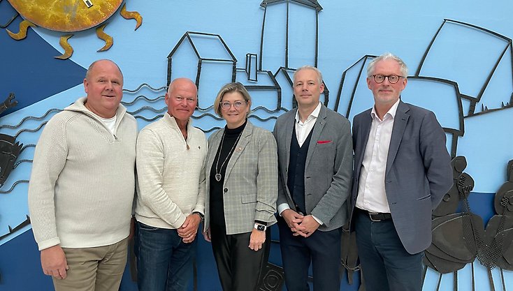 Fyra män och en kvinna i mitten står bredvid varandra och tittar in i kameran.