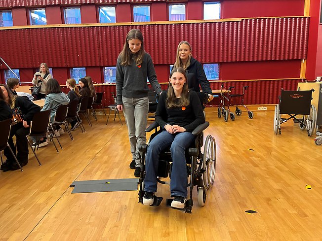 En tjej provar att sitta i en rullstol och köra över ett litet gupp. Bakom henne står två andra tjejer. 